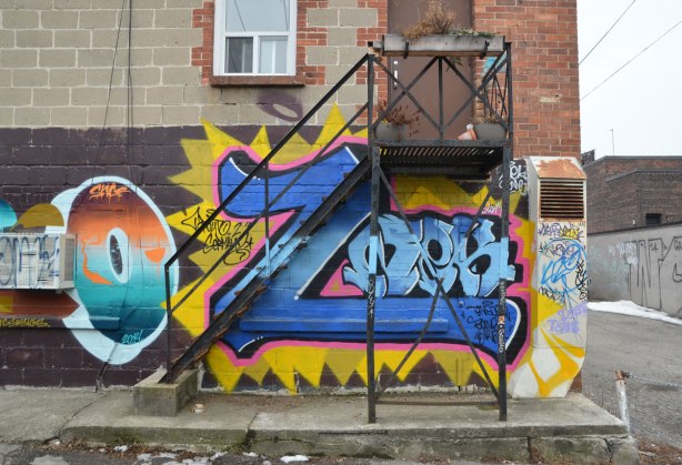 metal stairs to a small landing outside a door on the second storey of a concrete brick building.  The walls of the bottom floor are covered with graffiti.  Immediately behind the stairs is a large blue letter Z. 