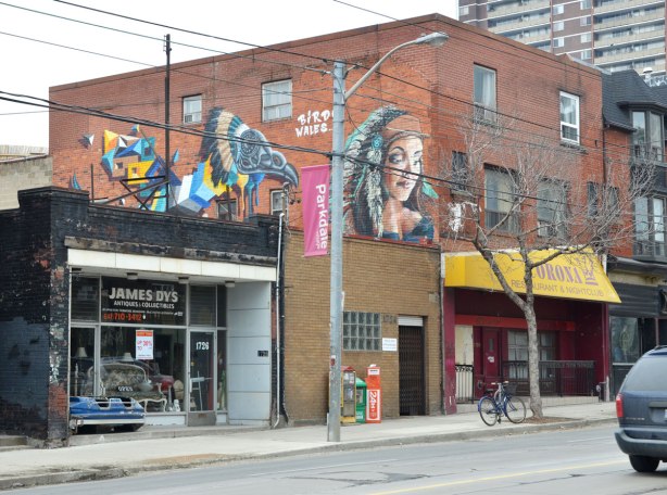 Two pieces of street art along the upper floor of a brick building.  In front, closest to the street is a young woman's head.  The other is a colourful stylized bird by the street artist Birdo.  The words "Birdo Wales" are written in white letter between the two pieces.  The Corona Restaurant with a yellow awning is on the right and James Dys Antiques and Collectibles is on the left.  is on the left. 