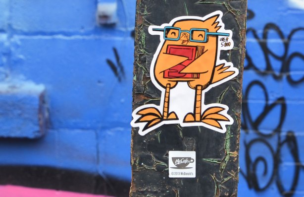 A small yellow bird on a small sticker.  It is an Uber 5000 bird, wearing glasses and holding a red book.  He's on a metal pole. 