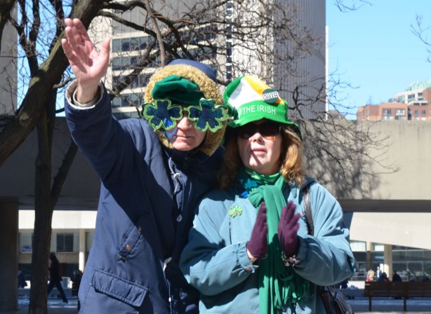 A couple watching a St. Patricks Day parade. He is wearing big shamrock shaped glasses. They are both wearing green hats. He is waving to the camera.
