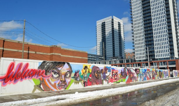 Colourful graffiti on hoardings around a construction site. Scenes of people and faces, kids on bikes, activities. The word Welcome is written in red cursive. Two apartment buildings are in thebackground.