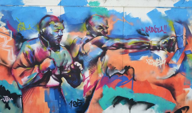 Colourful graffiti on hoardings around a construction site. Two men punching, one is punching while the other stands beside him with fists clenched