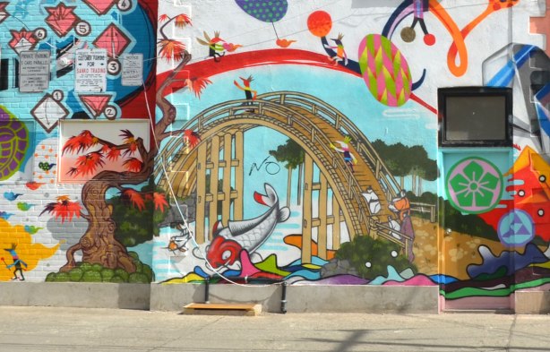 part of a mural on the side of a store on a corner lot that has a number of Japanese motifs.   Arched bridge over a stream with a fish jumping out of the water, Japanese maple tree beside the water, street art, large mural