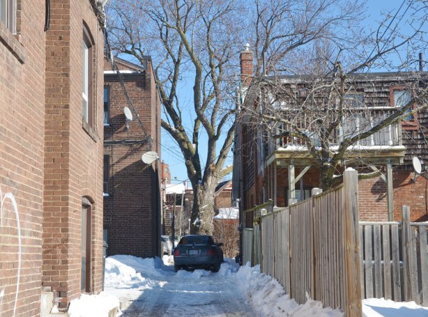 An alley with a wood fence on the right and a brick building on the left.  A car is parked at the end and just beyond the car is a large tree.  Winter time. snow. 