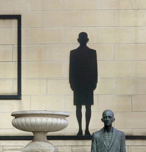 Close up of a bronze sculpture of a man.  In this picture you can only see his head and shoulders.  Behind him on a wall is a black silhouette that looks like his shadow.  A large white planter is also in the picture although there is nothing growing in it. 