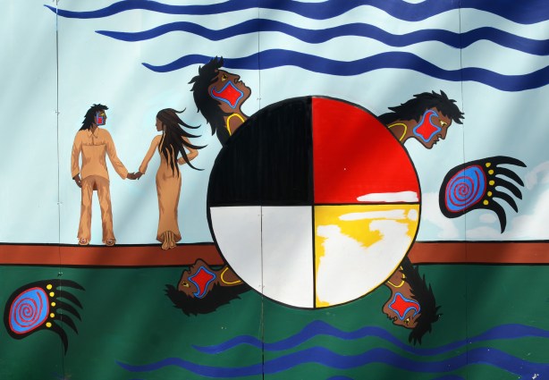 part of an Anishnawbe First Nations story/legend themed mural painted on wood construction hoardings in Allan Gardens - a couple walking hand in hand beside a circle divided into quarters, one is black and one is red and a head comes out of each quadrant. 