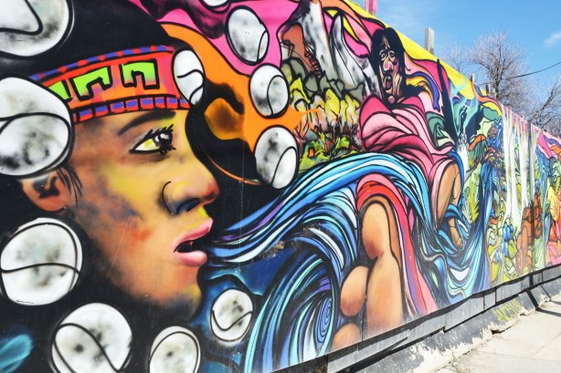 part of First Nations story/legend themed mural painted on wood construction hoardings in Allan Gardens, a young man's breath is stylized as blue ribbons streaming from his mouth 