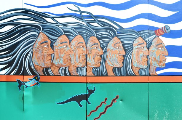 part of a First Nations story/legend themed mural painted on wood construction hoardings in Allan Gardens - a row of 6 men's heads seen in profile, all looking to the right, their long hair blowing away from their faces. 