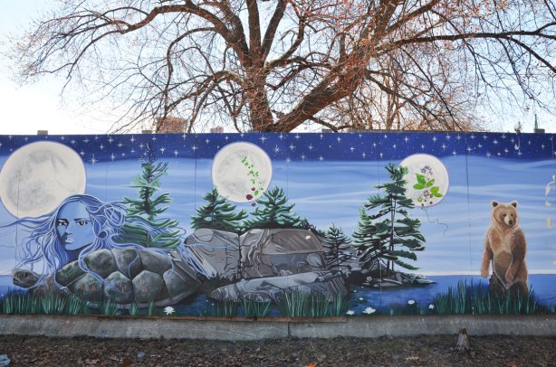 part of a First Nations story/legend themed mural painted on wood construction hoardings in Allan Gardens a leafless tree and a couple of smaller pines by a lake 