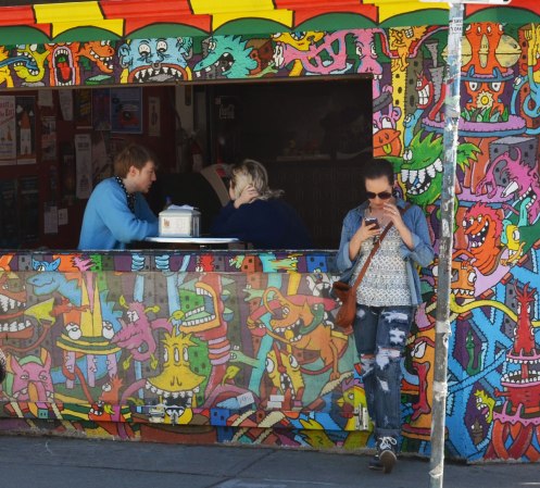 A couple is sitting by a window inside a restaurant.  The exterior wall of the building is covered in a mural of brightly coloured little monsters.  A woman is standing outside by the window as she checks her phone. 
