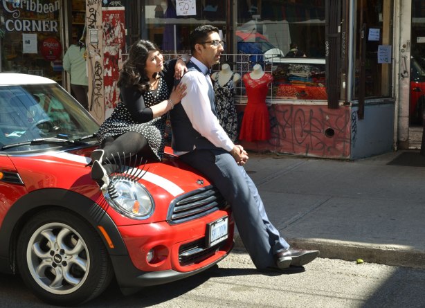 A couple is posing for engagement photos.  She is sitting on the hood of a red and white Mini Cooper.  He is leaning against the front of the car.  The car has big eyelashes over the front headlights.  The car is parked on a street in Kensington. 