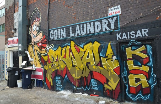 Graffiti and mural on the back of a coin laundry, spic and span laundry, and back entrance to Kaisar Guesthouse. Yellow and red tag with a picture of a guy wearing a red and white striped shirt.