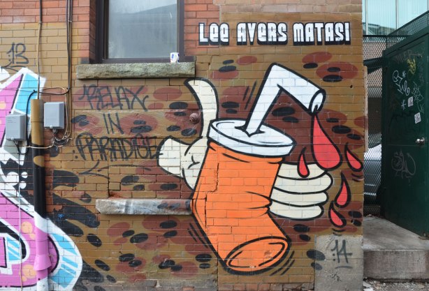 Picture of an orange pop can being crushed by a white hand. There are drops of red liquid (blood?) coming out of the straw that is in the top of the can. The words "Lee Avers Matasi" are written in block letters above the picture.