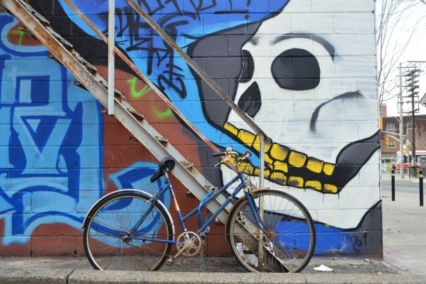 large white skull with yellow teeth, and mouth partially open painted on a wall by a metal exterior staircase. The skull is talking black symbols all jumbled up in a blue speech bubble. There is a bike parked in front of the stairs. Perhaps the skull is about to eat the bike?
