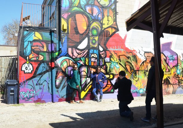 Two women are posing in front of a mural in a Kensington lane while a man takes a picture and another man watches. 