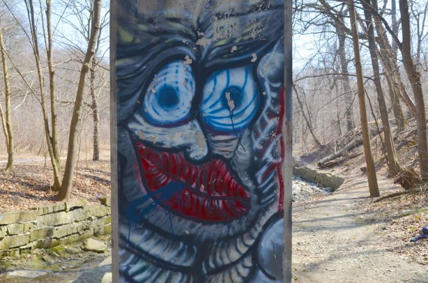 Graffiti faces on concrete pillars supporting a bridge - wild blue eyes and red lips with wavy skin, with the trees, creek and path through the ravine in the background. 