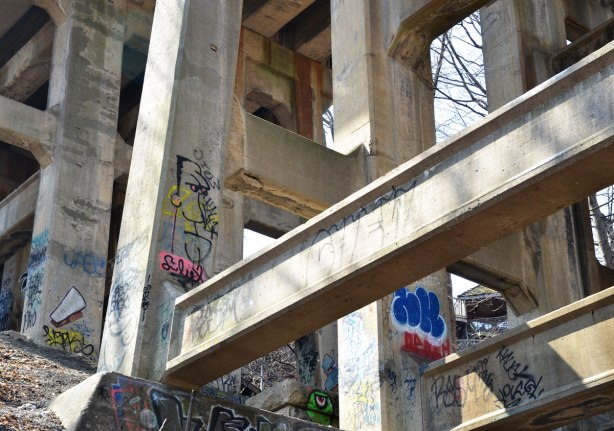 Graffiti faces on concrete pillars supporting a bridge - looking up from below so there are a large number of supports visible.  A line drawing of a man's head is on one of the pillars