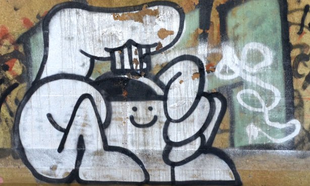 Graffiti faces on concrete pillars supporting a bridge - the smiley face is on a spray paint can, a large finger is on the trigger. 