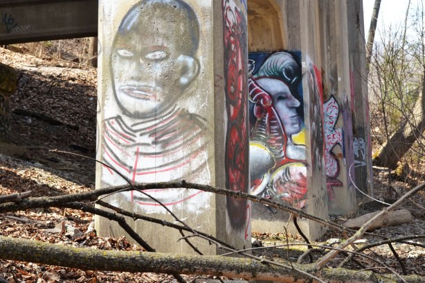 Graffiti faces on concrete pillars supporting a bridge - three supports in a row are visible.  In the foreground is a simple drawing of a man's head in blacks and greys, the next pillar has a more elaborate man in blues 