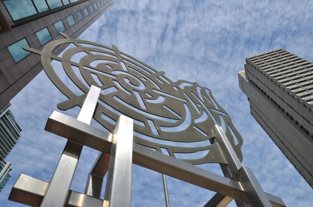 Looking upward from below at a public art installation called 'Nautilius' by Judith Scwarz which is a two dimensional cut out piece of metal of a snail-like spiral, mounted about ten to 12 feet off the ground on square sided metal poles.