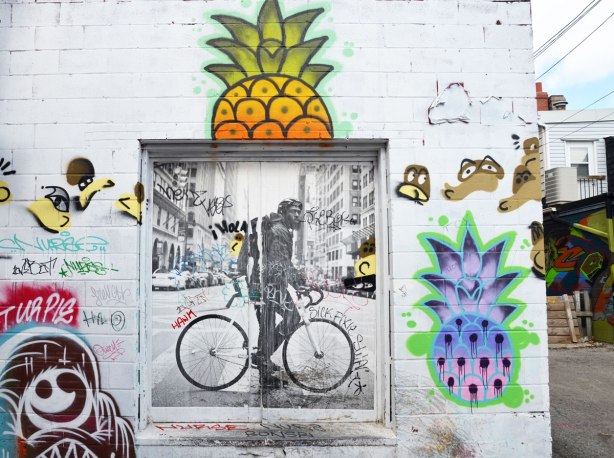 Square wheatpaste graffiti of a man on a bicycle on a city street. Two large pineapples have been painted on the wall too, one above the bicycle man and one to the right. The pineapples are bright colours, one is orange and yellow and the other is blue and purple.