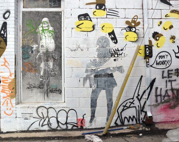 Wheatpaste and paper graffiti on a white concrete block wall. A pair of yip yaps beside a person with an owl head and one wing instead of an arm. The second arm is normal. Also in the picture is a heatpaste black and white picture of a woman riding a bicycle away from the viewer. She is wearing polka dot shorts. Someone has also painted yellow and black bee like creatures with faces (black masks) and brown hats.