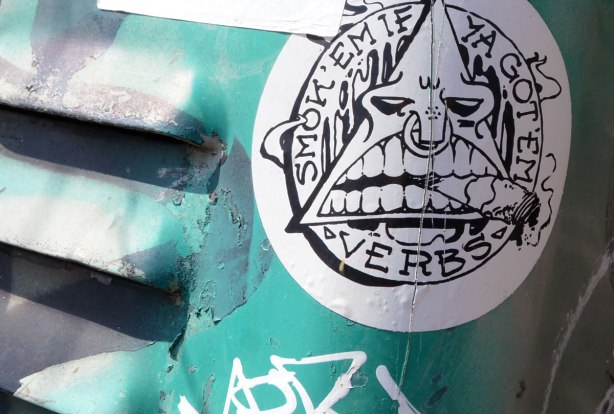 Sticker on a teal coloured metal box.  Sticker has triangle inside a circle.  The triangle is made into a face, big open mouth with cigarette clenched between the teeth.  Around the circle are the words 'Smok'em if ya got 'em. Verbs