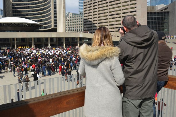 a couple taking photos from the upper level of Nathan Phillips square.  Down in the square below them is a large pillow fight, and a lot of spectators and photographers