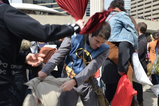 A young man in a batman shirt is being hit over the head with a red pillow in a large pillow fight. 
