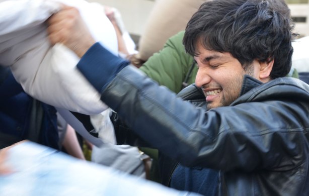 Young man in a black leather jacket has his eyes closed as he hits someone with a pillow in pillow fight. 