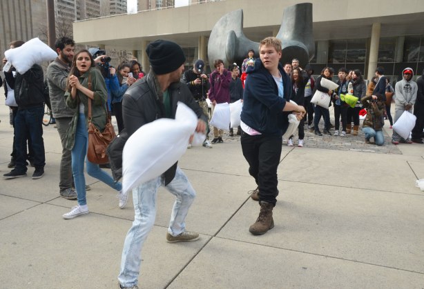 Two young men having a pillow fight while a crowd stands around them in a circle and watches. 