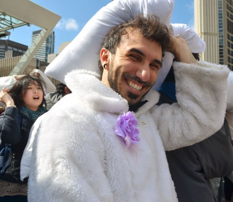 Man with a beard and a moustache, wearing a white fuzzy jacket with a pale purple artifical flower pinned to it. He's smiling and holding a pillow over his head. 