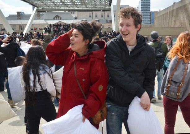 A happy couple taking a break from a pillow fight.  He is laughing, she is saying ouch and putting her hand over her ear. 