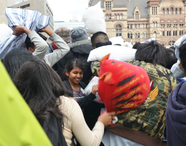 A young woman has her eyes closed and is smiling as the people around her are hitting each other with pillows. 