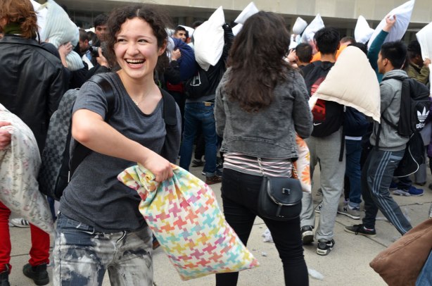 A young woman is holding a pillow with a pillowcase that has pink, green and yellow geometric shapes on it.  She is getting ready to hit someone with it in a pillow fight.  She is laughing  