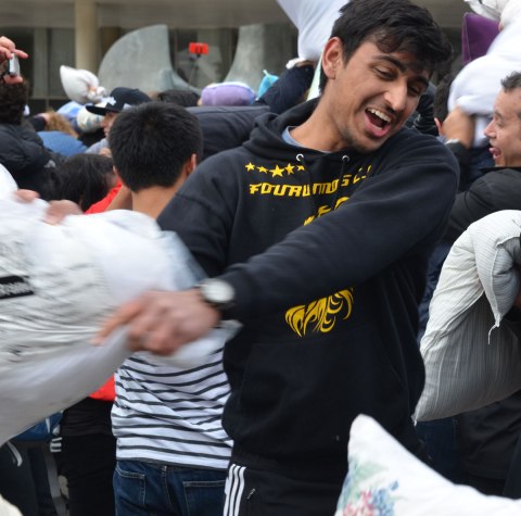 A young man is swinging a pillow while someone on the left is taking his photo. 
