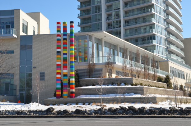 Looking across the street (Don Mills Rd) at a new condo development.  Three tall striped poles are beside one of the buildings as part of an art installation.  The building closest to the poles is low rise (2 or 3 storeys) but the building behind is a much taller structure. 