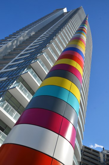 A brightly coloured striped cylindrical shaped pole in front of a tall condo building. 