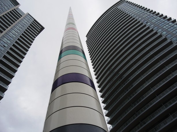 Very tall striped cylindrical pole with mostly white stripes with a few greys and only one or two pale colours.   Looking up from the base of it towards the tip, two tall condos, one on either side, are in the photo too. 
