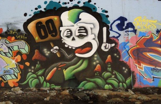 part of a mural under a bridge - a wacky white face with open mouth.  One hand of the creature is holding a sign that says 69 and the other hand has one finger pointed upwards