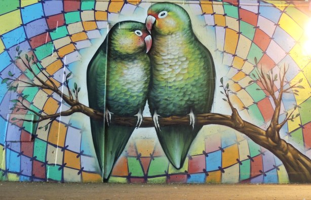part of a mural on a concrete wall under a bridge under the 401 in Toronto, two green lovebirds on a tree branch, with a background of multicoloured diamond shapes arranged concentrically 