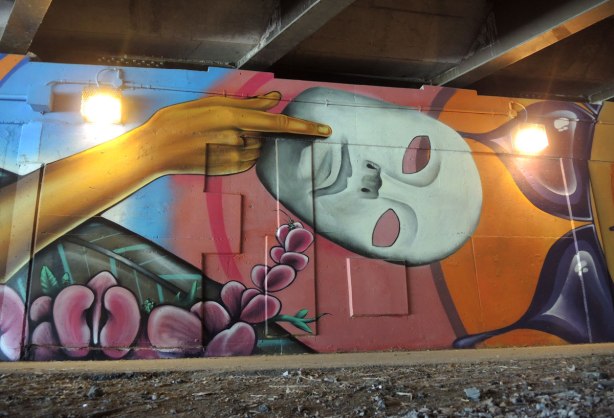part of a mural on a concrete wall under a bridge under the 401 in Toronto, a hand is holding a white mask. 