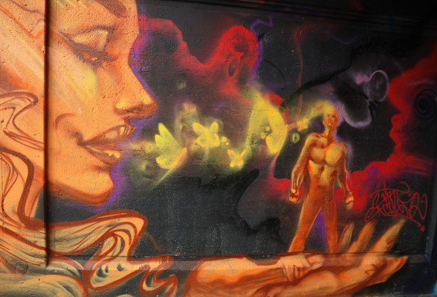 street art painting under a railway bridge - a large woman's face, she is breathing on a little man who is standing on her outstretched hand. 