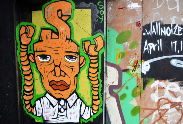 street art painting under a railway bridge - a mn with orange skin, large head and twisted pipes for arms.  A bis S is coming out of the top of his head