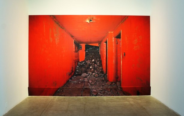 A large photograph of the inside of an abandoned building.  The interior walls have been painted red.  Rubble has started spilling into the hall from the demolition of the rest of the building. 