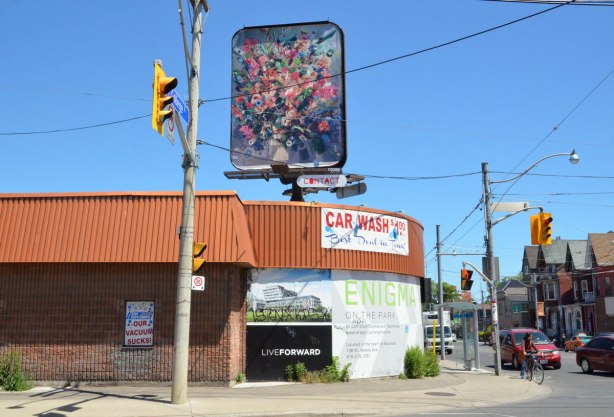 An building with a 'car wash' sign on it but the windows are covered with a sign for "engima' cond development.  On the roof of the building is a billboard with a large photo of a flower arrangement. 