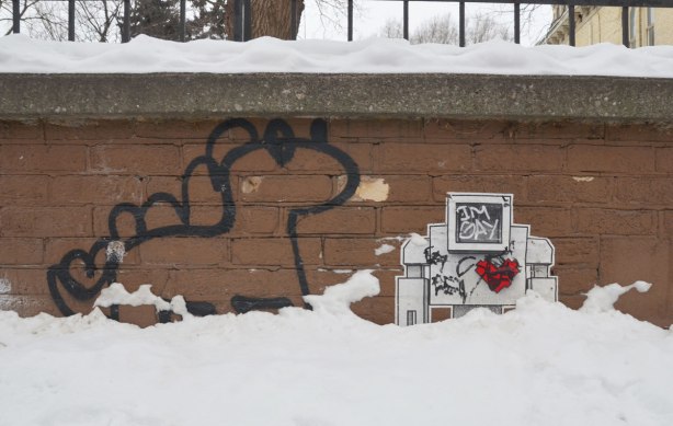 lovebot on a low brick wall, in the snow. A black line drawn dinosaur is beside him. Someone has scribbled 'I'm gay' on the lovebot