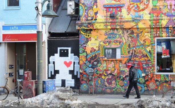A painted lovebot about person height on a wall beside Lee's Palace on Bloor St - a building that is covered with intricate bright coloured pictures