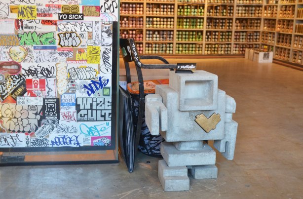 a little cement 3D lovebot statue stands on the floor of a store that sells cans of spray paint