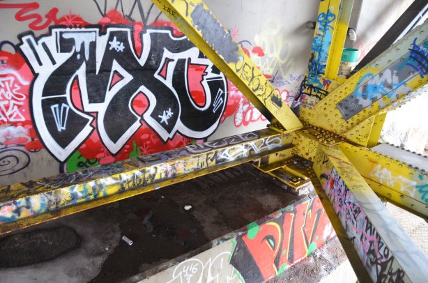 graffiti on the yellow girders under the bridge. The concrete supports at the end of the bridge are also visible and also covered with graffit.